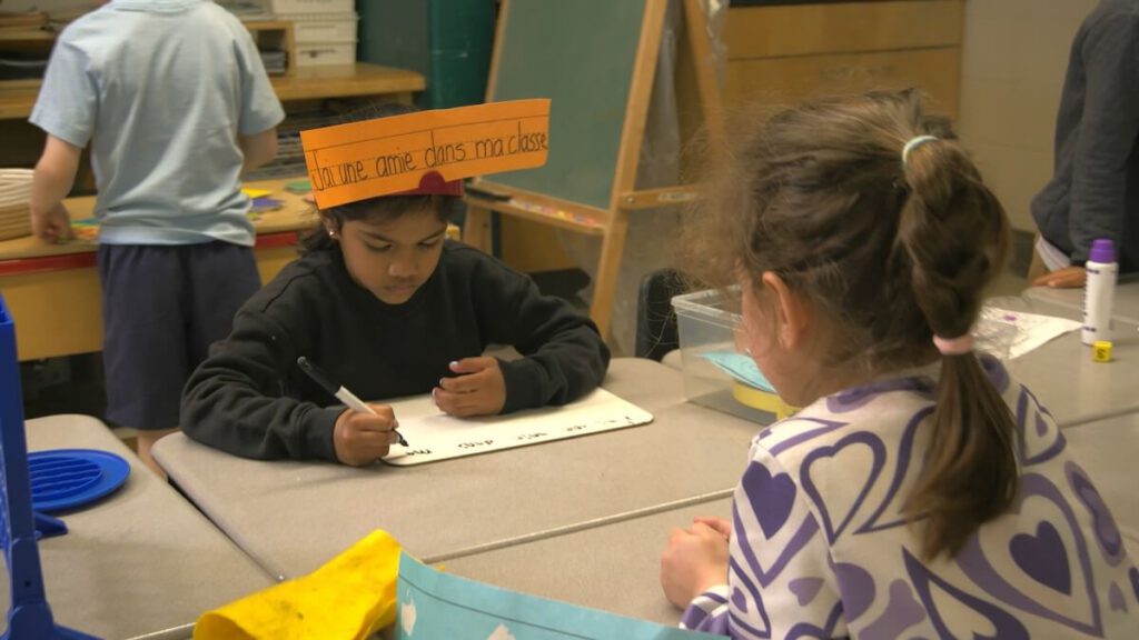 A girl is seated at a classroom desk and has a piece of orange paper on her head with some words for a vocabulary exercise.